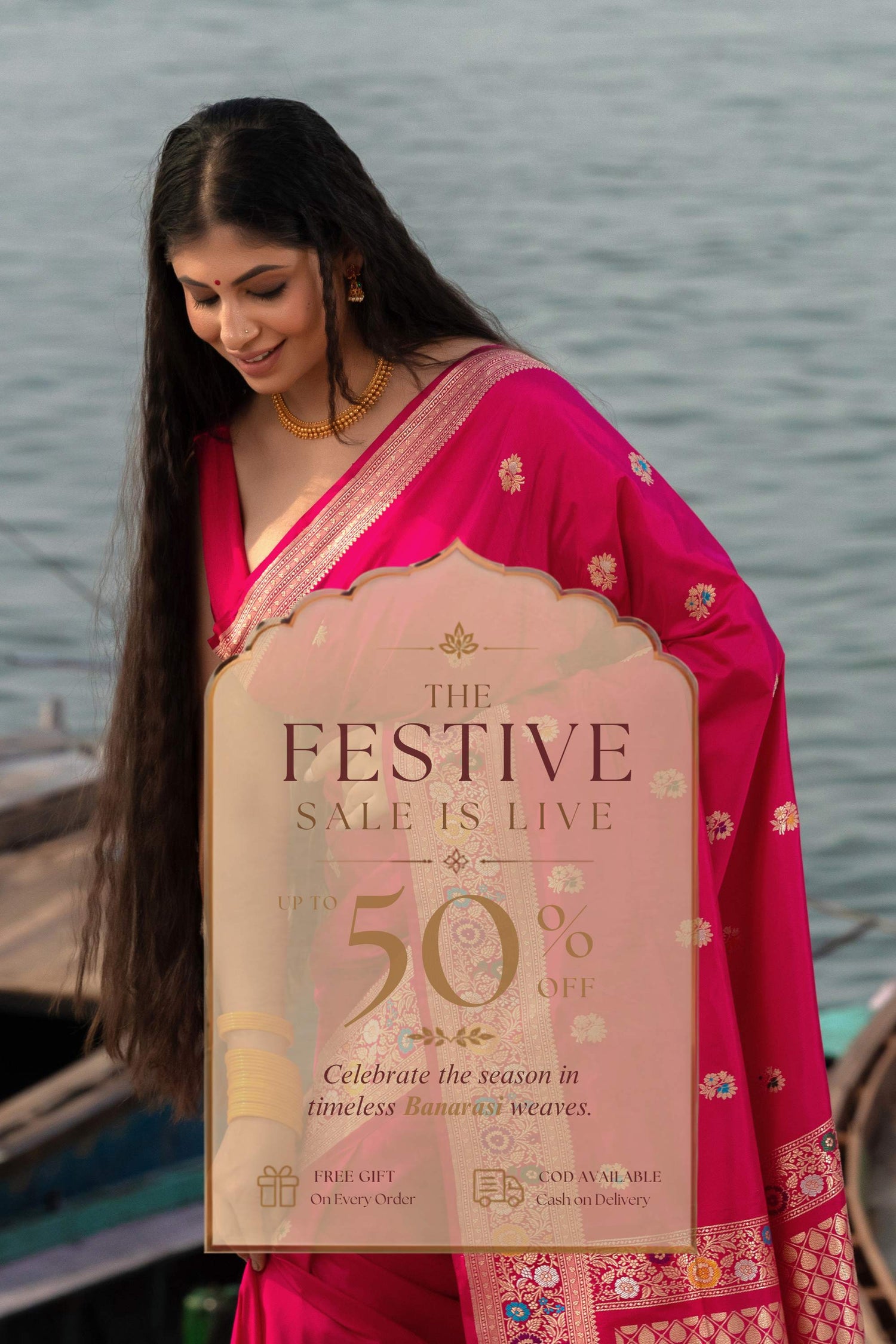 A woman wearing a bright pink saree stands outdoors near water, holding a large promotional sign that reads 'The Festive Sale Is Live' and '50% Off'.