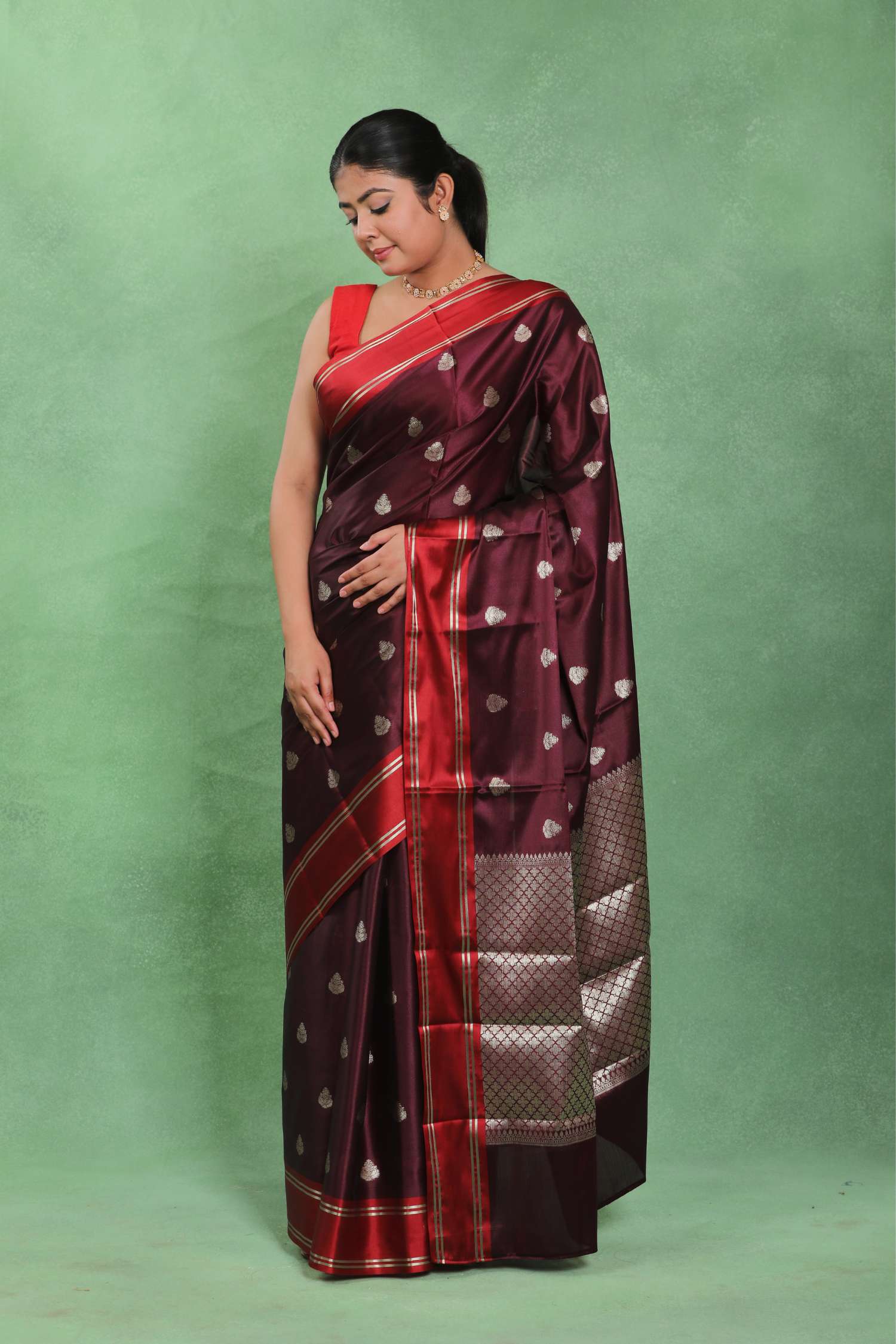 A woman modeling a dark maroon silk saree with red and gold borders, featuring small metallic heart motifs, set against a solid green backdrop.
