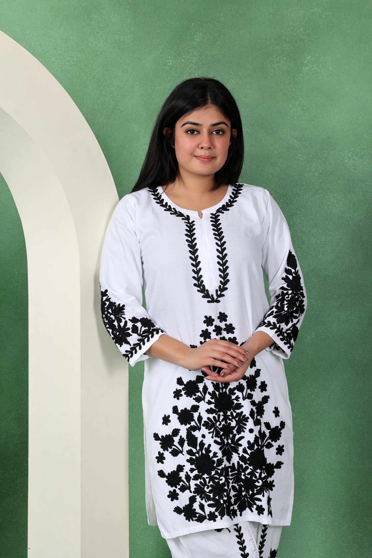 A woman modeling a white kurta with black floral embroidery, standing against a textured green backdrop with a white arched prop on the left.