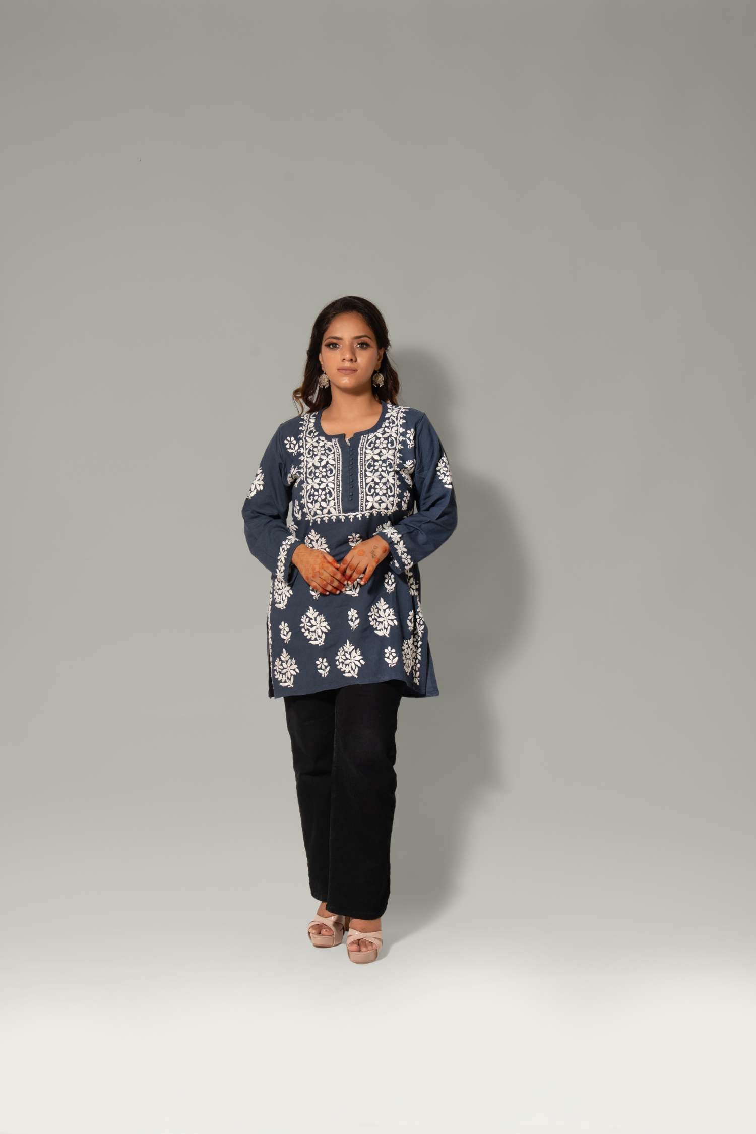 A woman modeling a navy blue tunic top with intricate white embroidery, paired with black pants, standing against a neutral gray studio background.