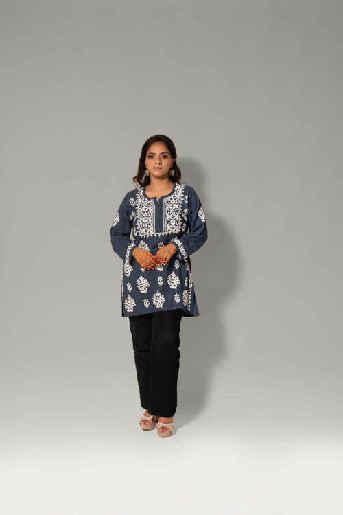 A woman modeling a navy blue tunic top with intricate white embroidery, paired with black pants, standing against a neutral gray studio background.
