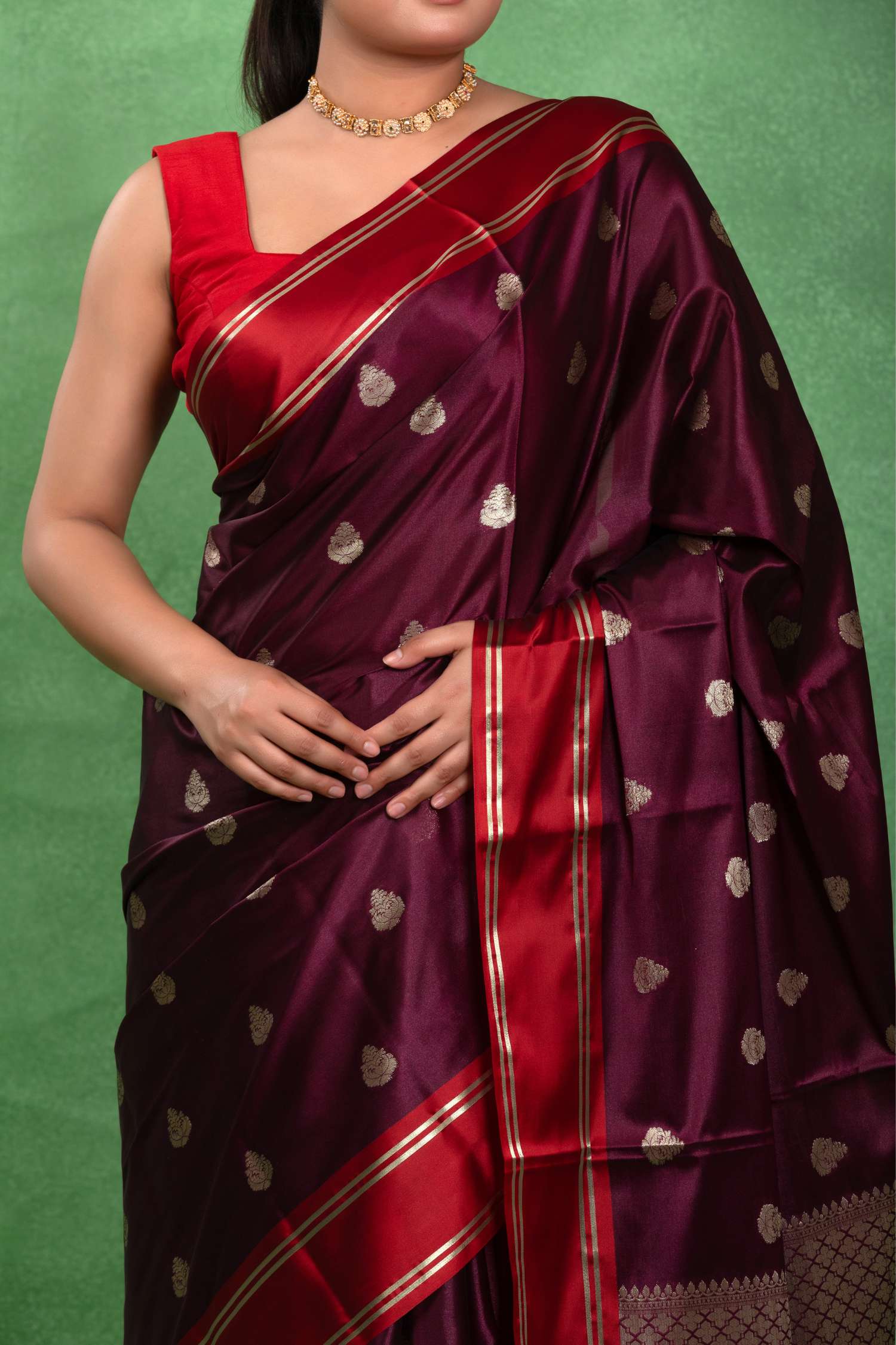 A woman wearing a deep maroon silk saree with red border stripes and gold zari motifs, paired with a matching red blouse, set against a textured green backdrop.