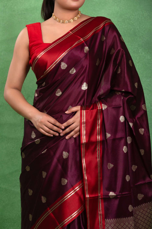 A woman wearing a deep maroon silk saree with red border stripes and gold zari motifs, paired with a matching red blouse, set against a textured green backdrop.
