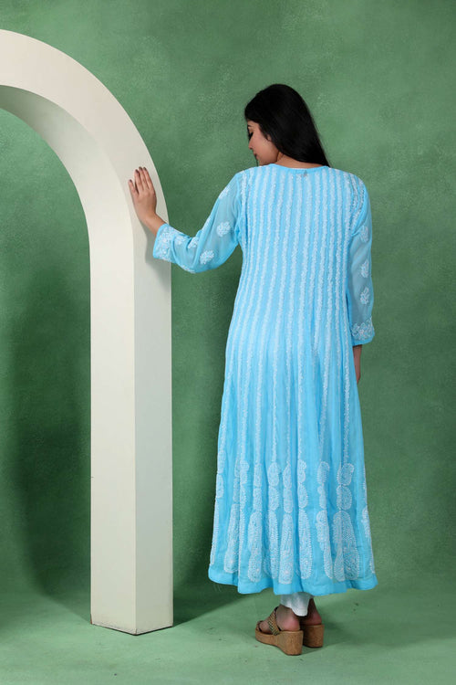 A woman wearing a light blue and white vertically striped long kurta stands with her back to the camera, touching a white arched structure against a textured green studio backdrop.