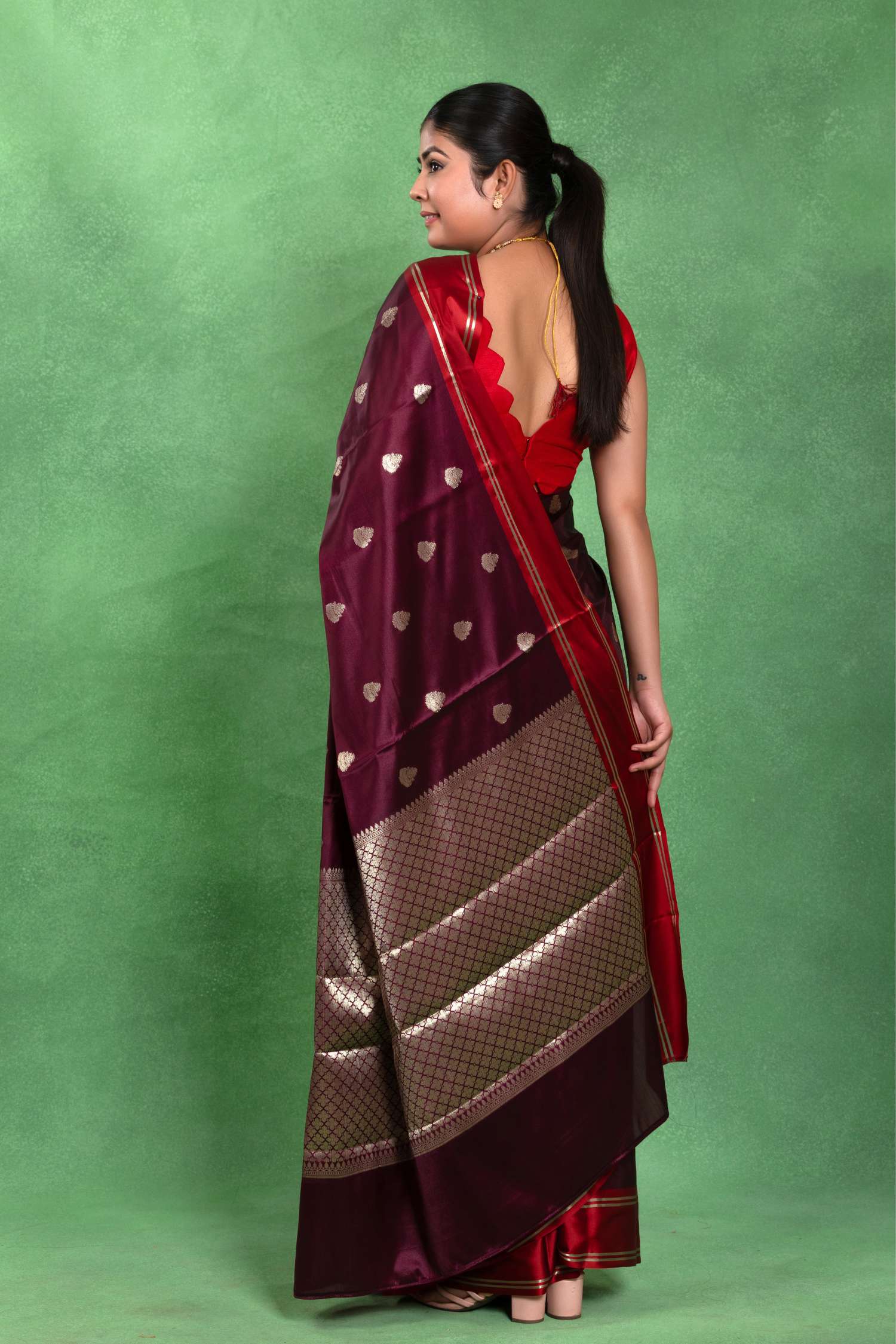 A woman modeling a deep maroon silk saree with metallic gold zari patterns, a gold border, and a red scalloped neckline blouse, posed against a solid green backdrop.
