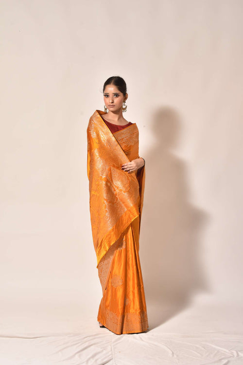 A woman modeling a golden-orange silk saree with subtle metallic sheen and a deep maroon blouse, standing against a plain beige backdrop.