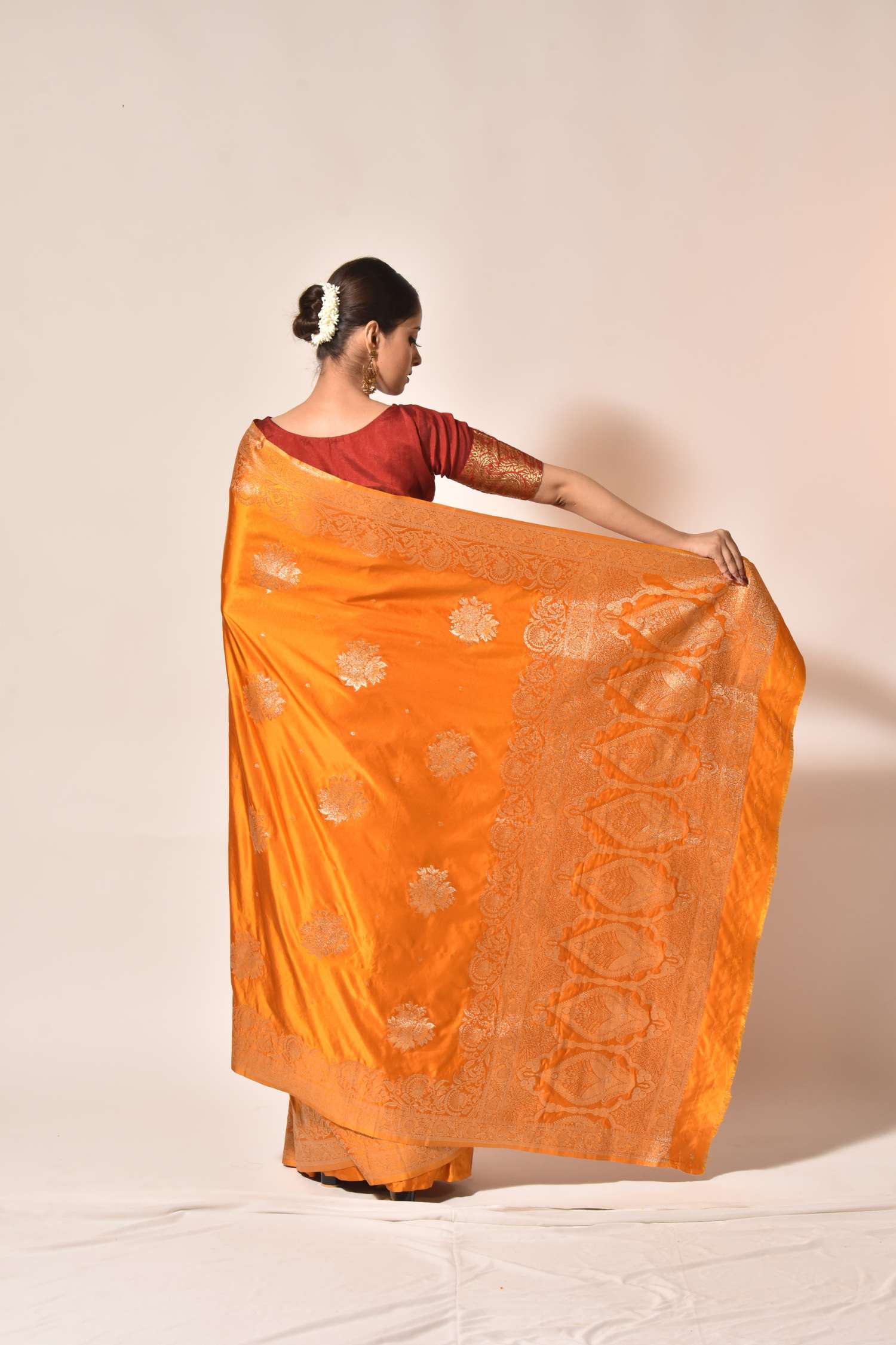 A woman wearing a bright orange silk saree with gold zari motifs and a red blouse, holding the saree out to display its drape and pallu against a plain beige studio background.