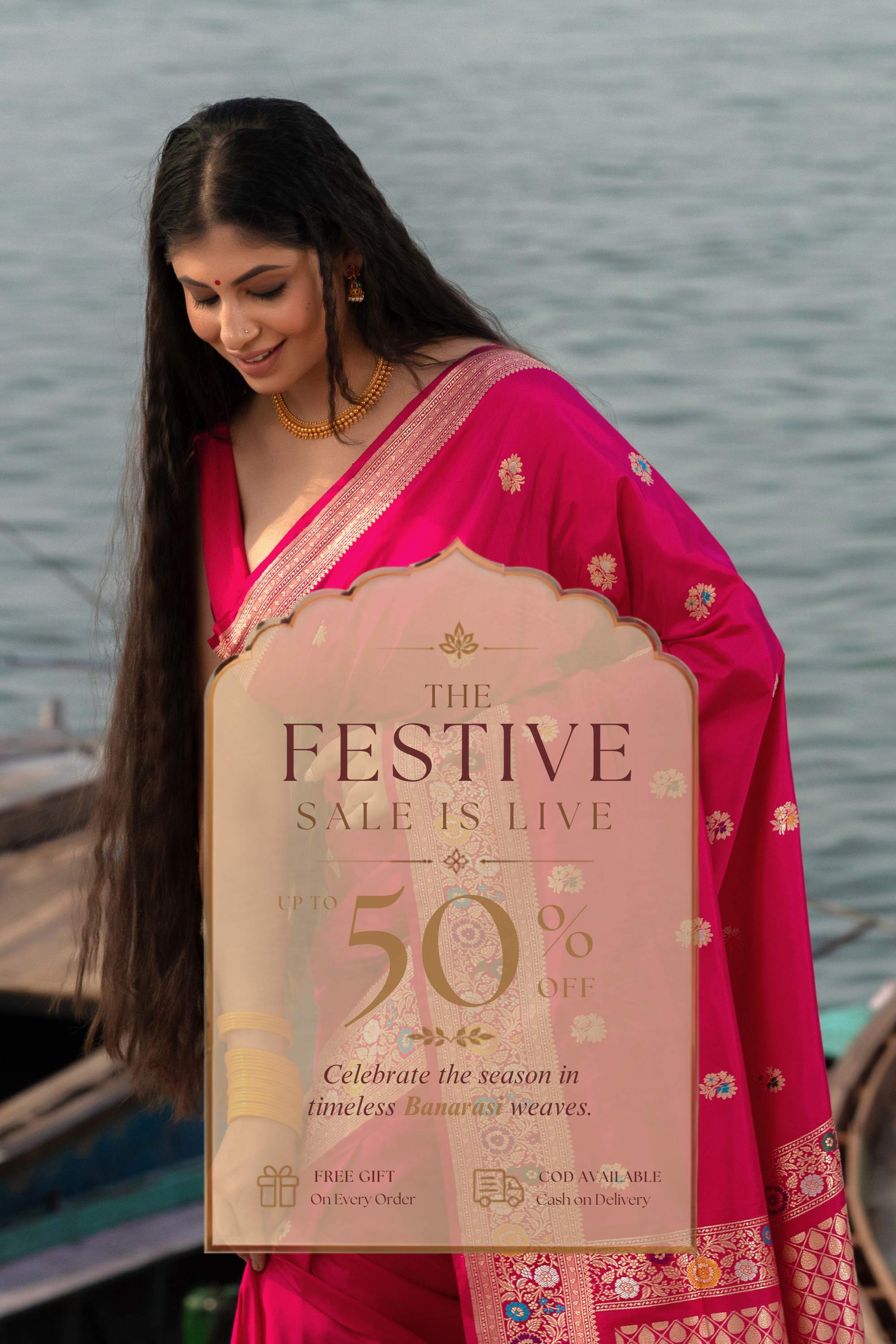 A woman wearing a bright pink saree stands outdoors near water, holding a large promotional sign that reads 'The Festive Sale Is Live' and '50% Off'.