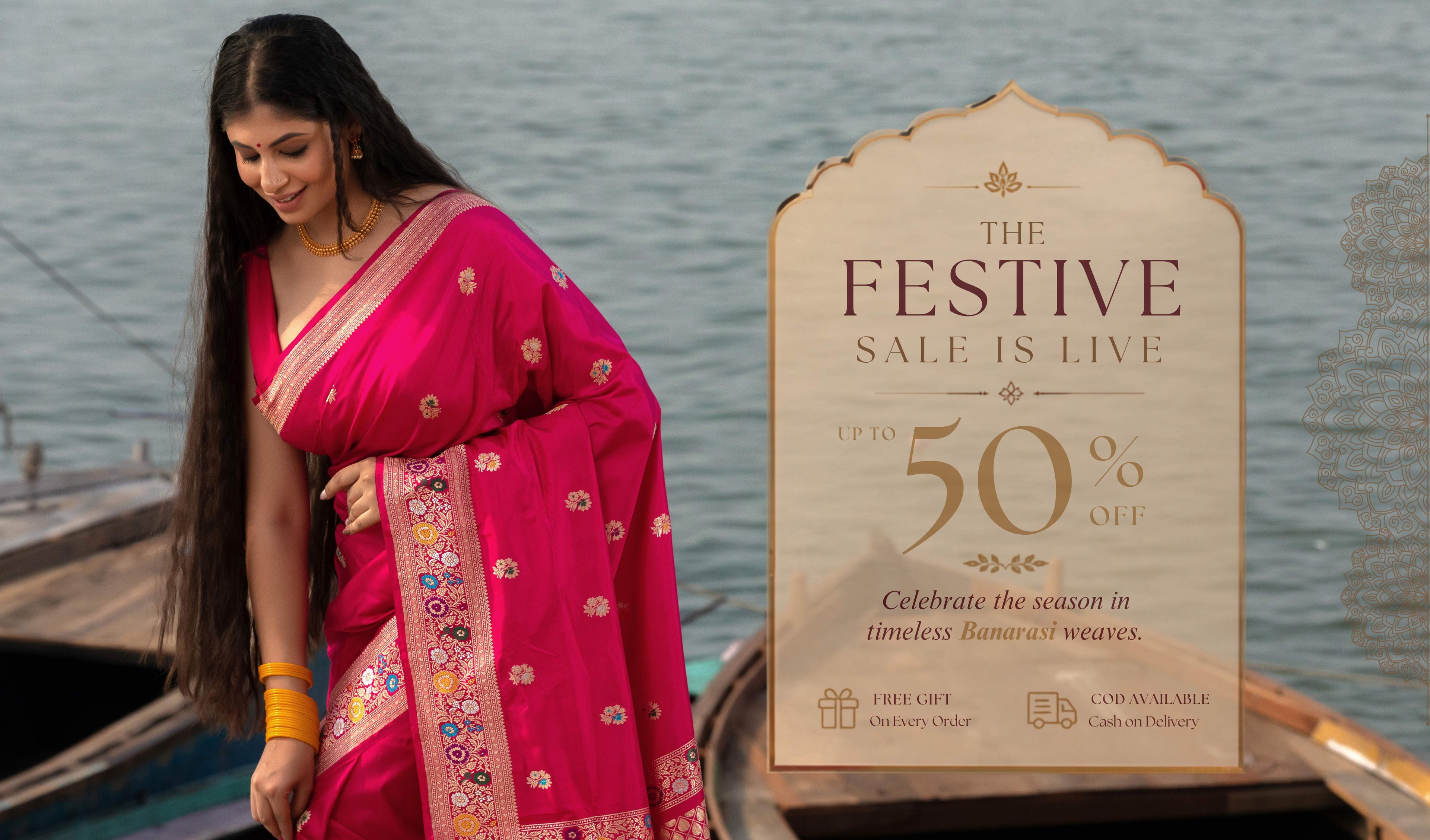 A woman wearing a bright pink Banarasi saree with gold and multicolor floral embroidery stands next to a large promotional sale sign that reads 'The Festive Sale Is Live Up To 50% Off'. The background shows a scenic view of water and boats.